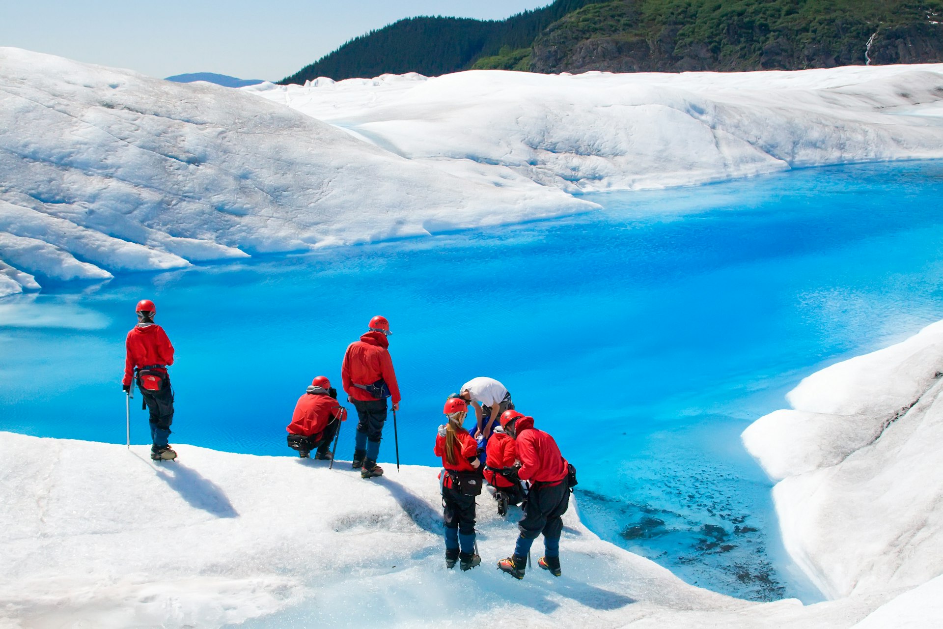 A group of trekkers on a glacier wearing bright red suits gaze down upon a turquoise icy lake