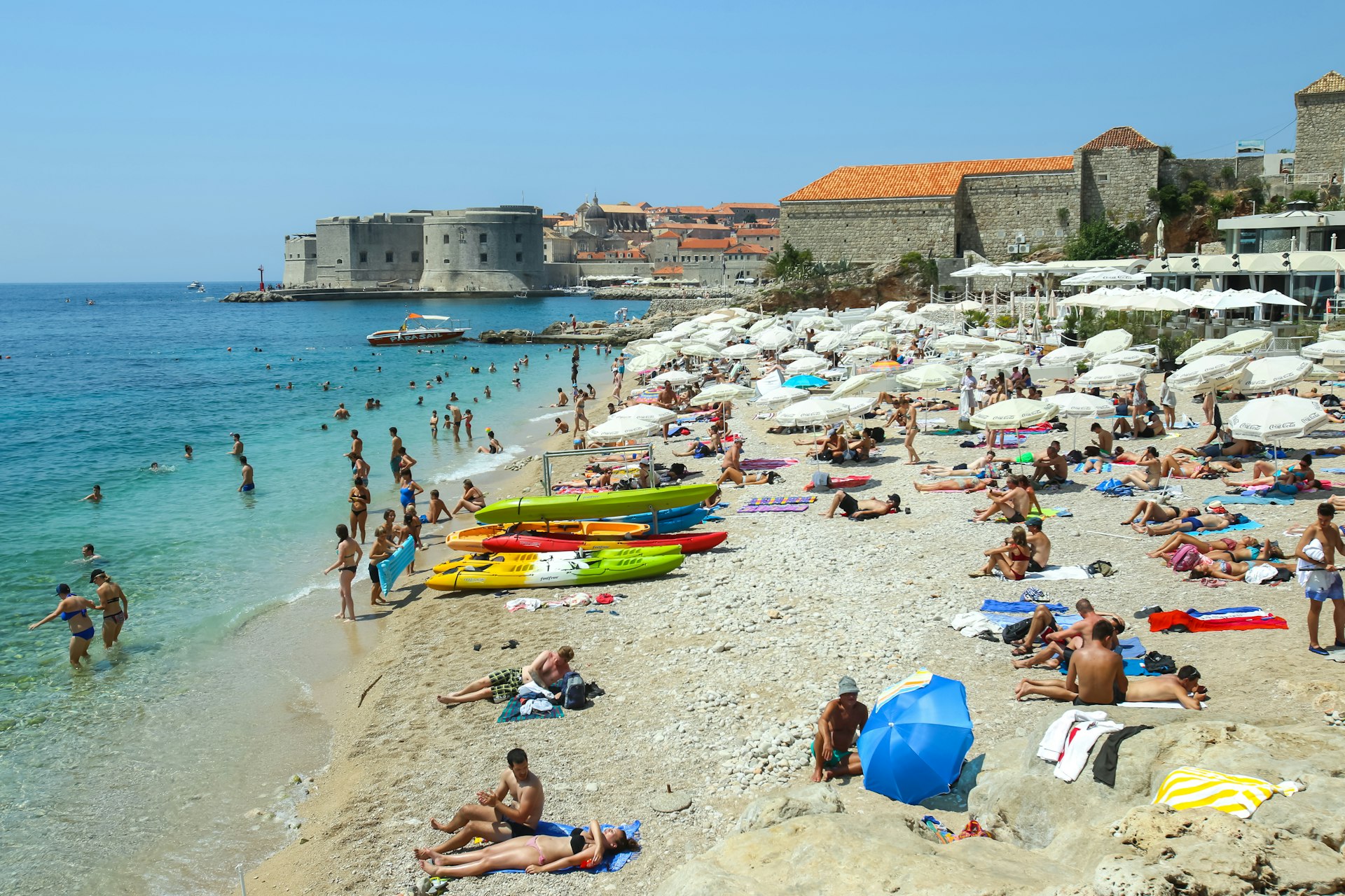 People swimming and sunbathing on the Banje sea beach, with the old town of Dubrovnik in the background.