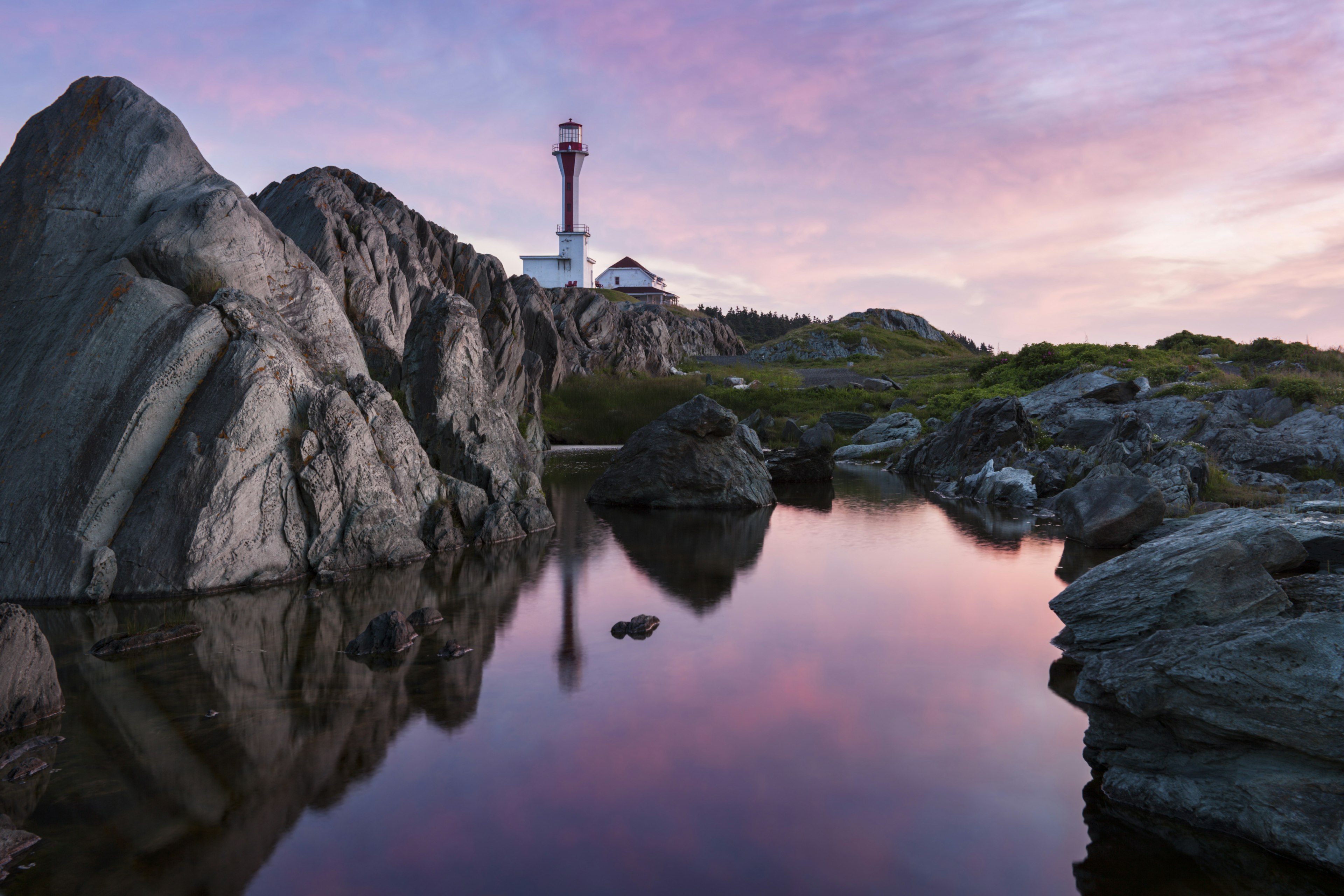 A rocky shoreline at sunrise with a purple-pink sky reflected in the water and Cape Forchu lighthouse in the distance