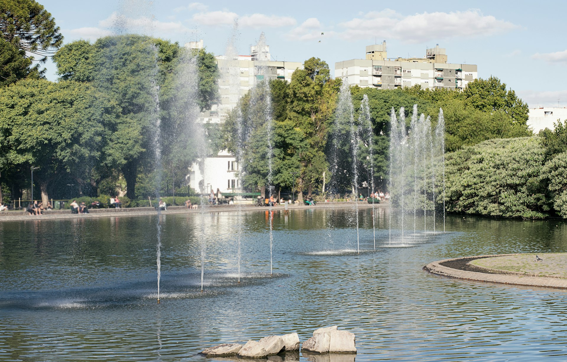 A large artificial lake with fountains; people are milling around in the surrounding park