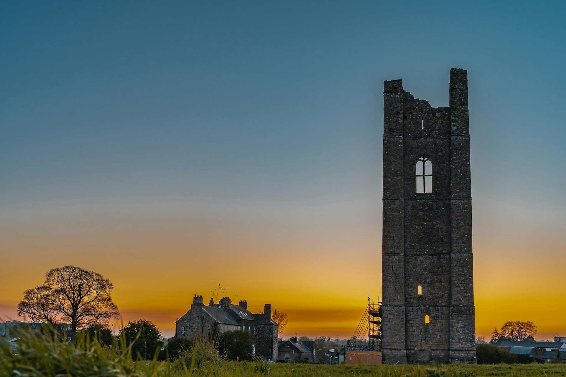 The sun sets behind a tall ruined stone Gothic tower in a rural area