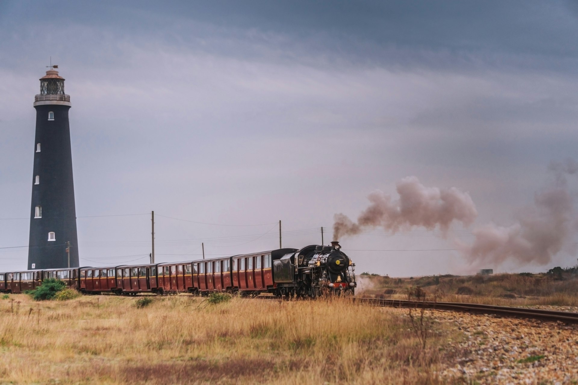 A tall free-standing lighthouse with black bricks is passed by a steam train with red carriages