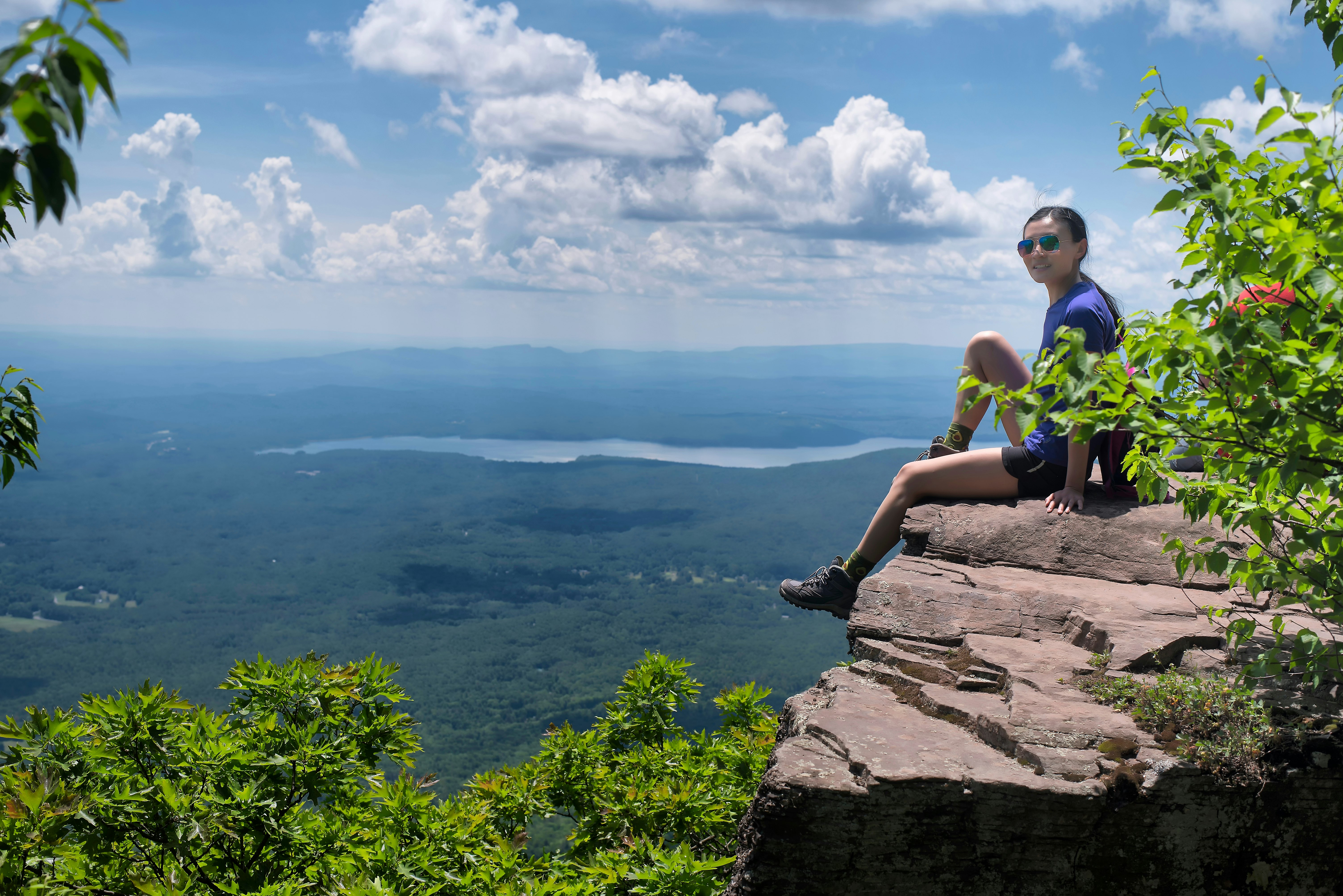 A chinese woman sitting on the edge of a cliff on top of overlook mountain in the catskills in Woodstock New York on a sunny summer day.;