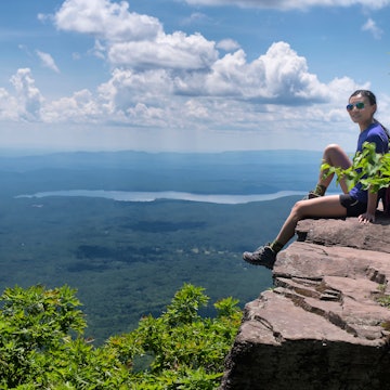A chinese woman sitting on the edge of a cliff on top of overlook mountain in the catskills in Woodstock New York on a sunny summer day.;