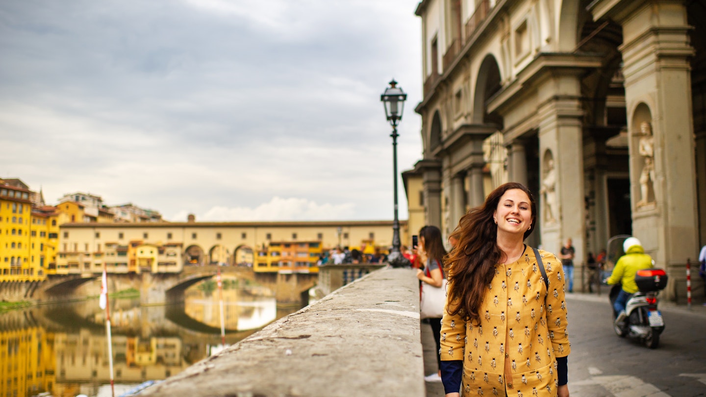 A tourist girl with an orange coat happily walks on the embankment in Florence, Italy.
