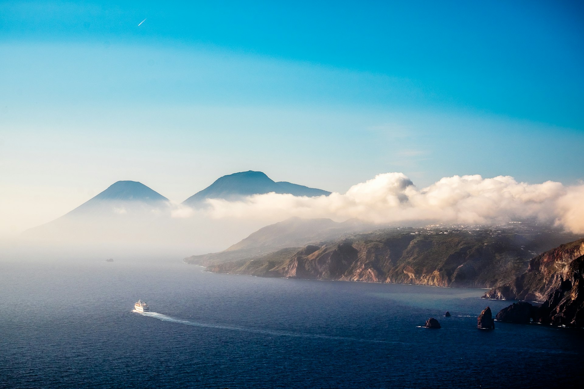 A ferry leaves a long curve of white water in its wake as it travels between craggy. mist-covered volcanic islands