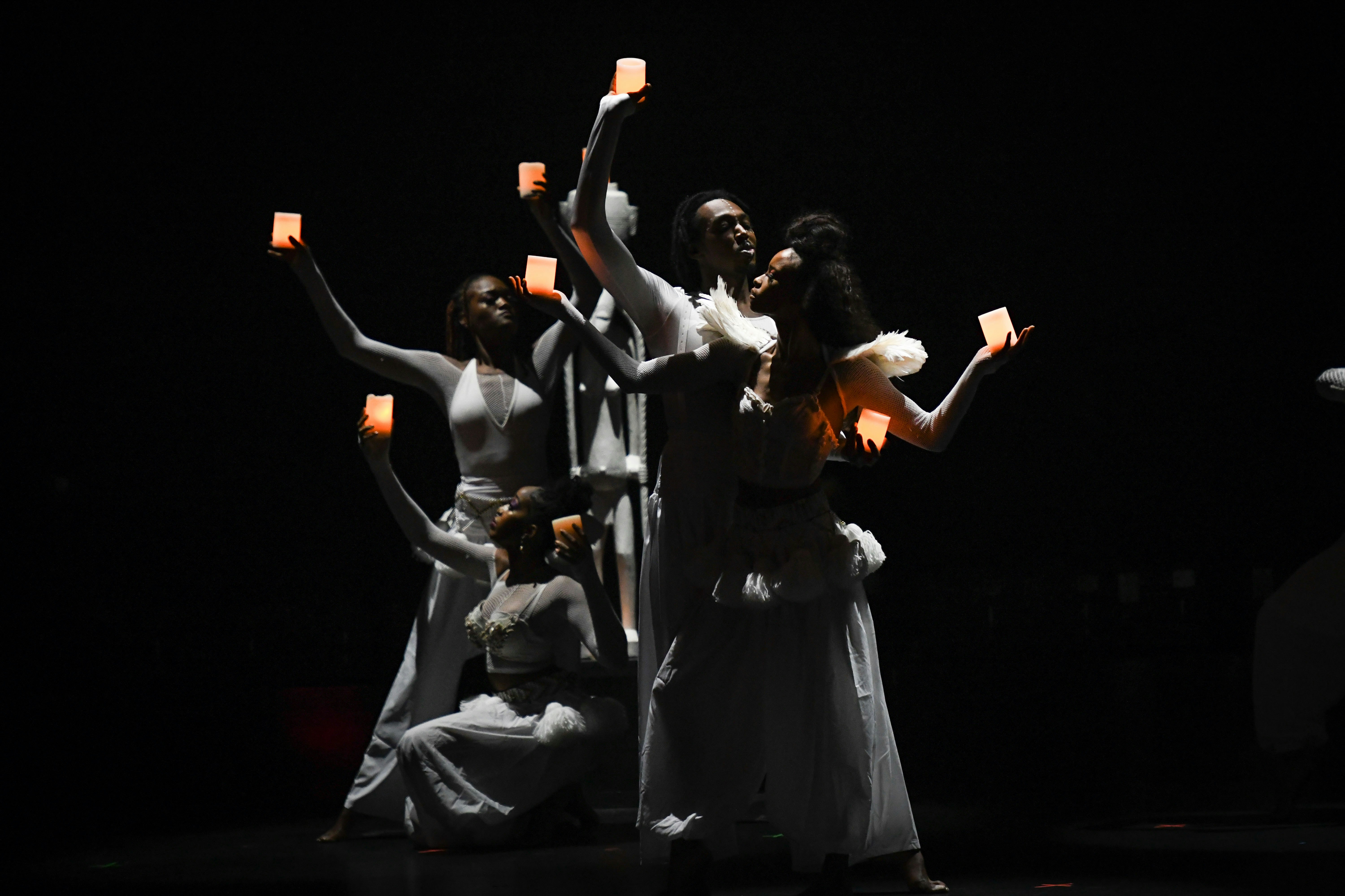 Black dancers dressed in white, posed in front of a dark backdrop and holding candles aloft