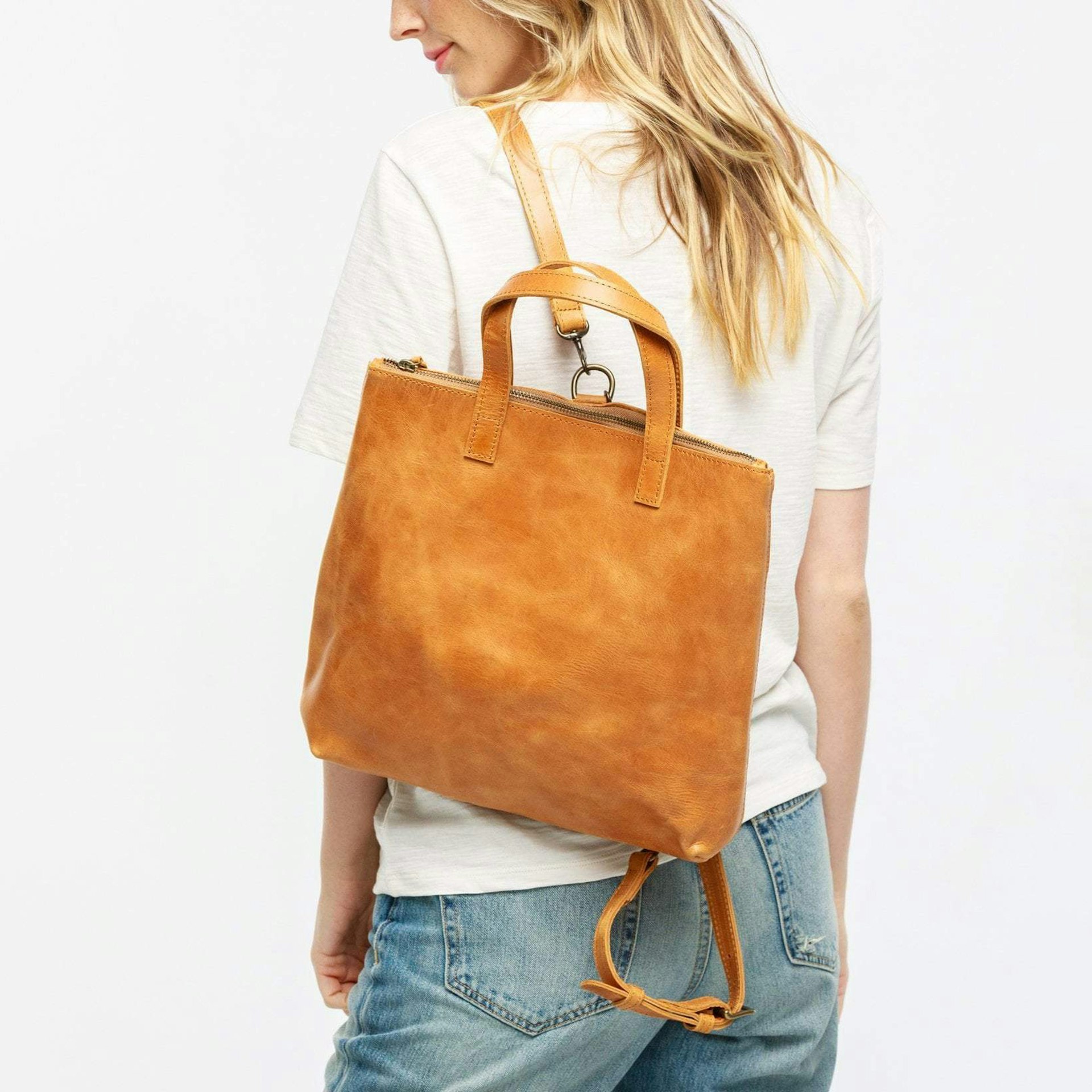 A blond woman in a white shirt seen from the back, wearing Able's Abera convertible backpack in camel over one shoulder