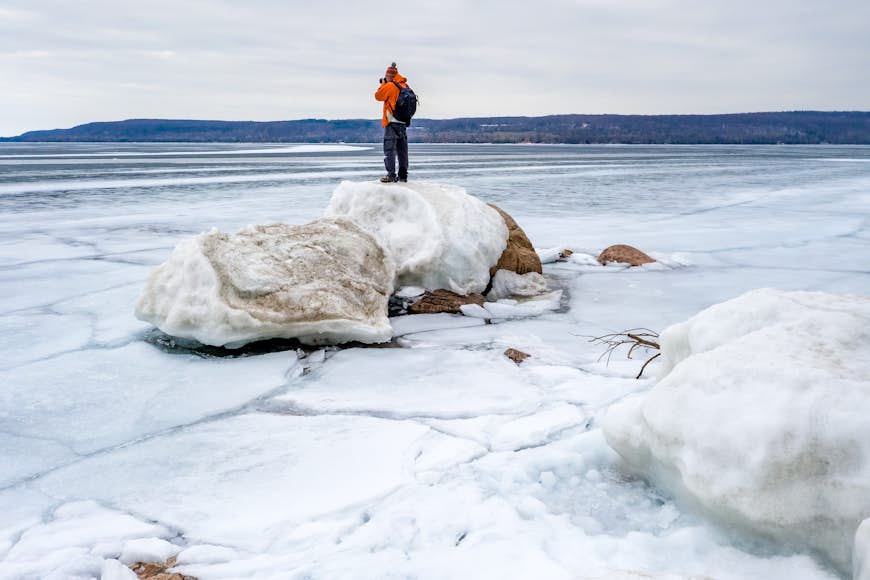 A man stands on a snow-covered rock taking a picture of the frozen bay in Ontario
