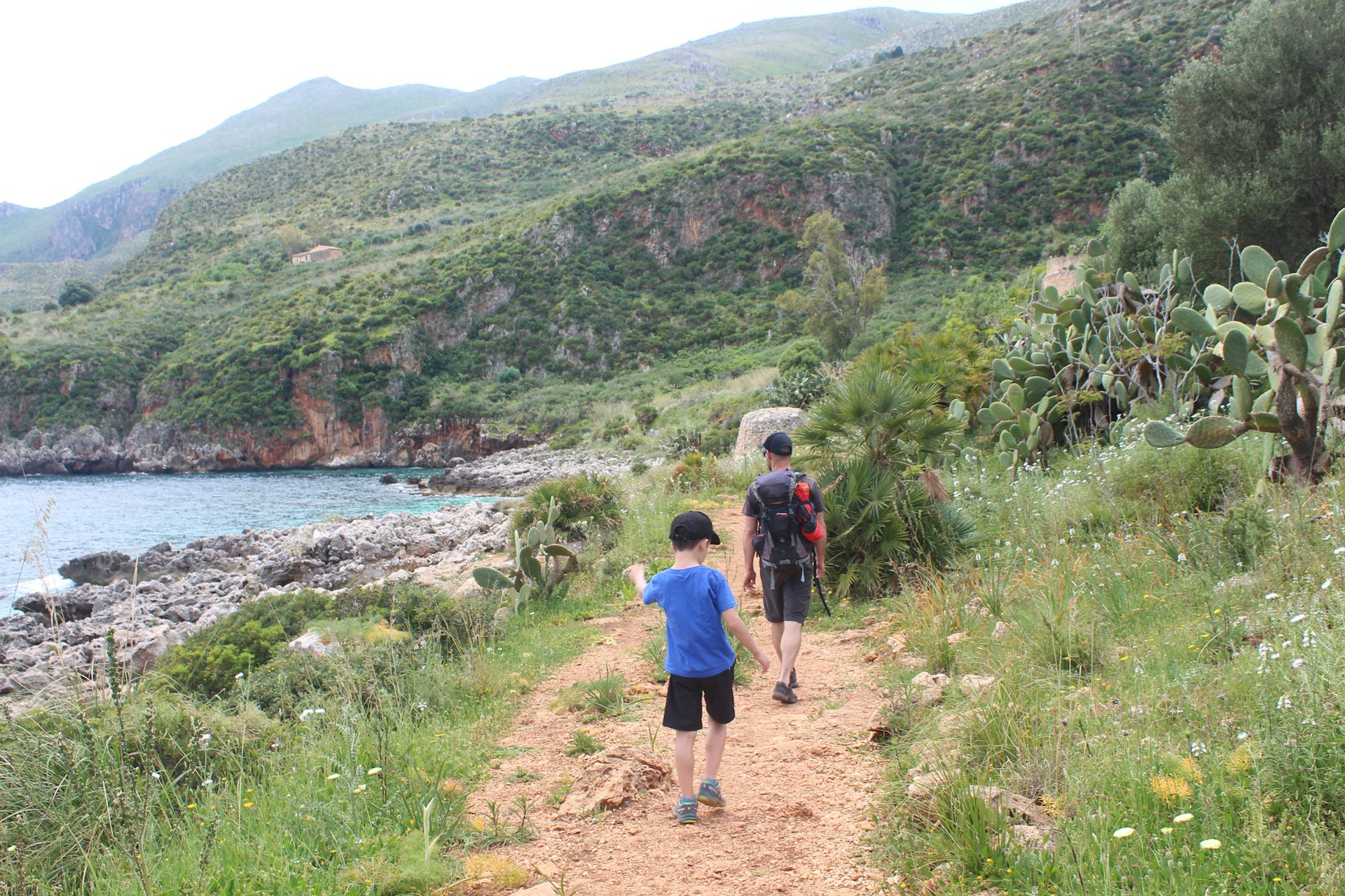 An adult and a child walk away from the camera along a path. The sea is to their left; foliage is to their right