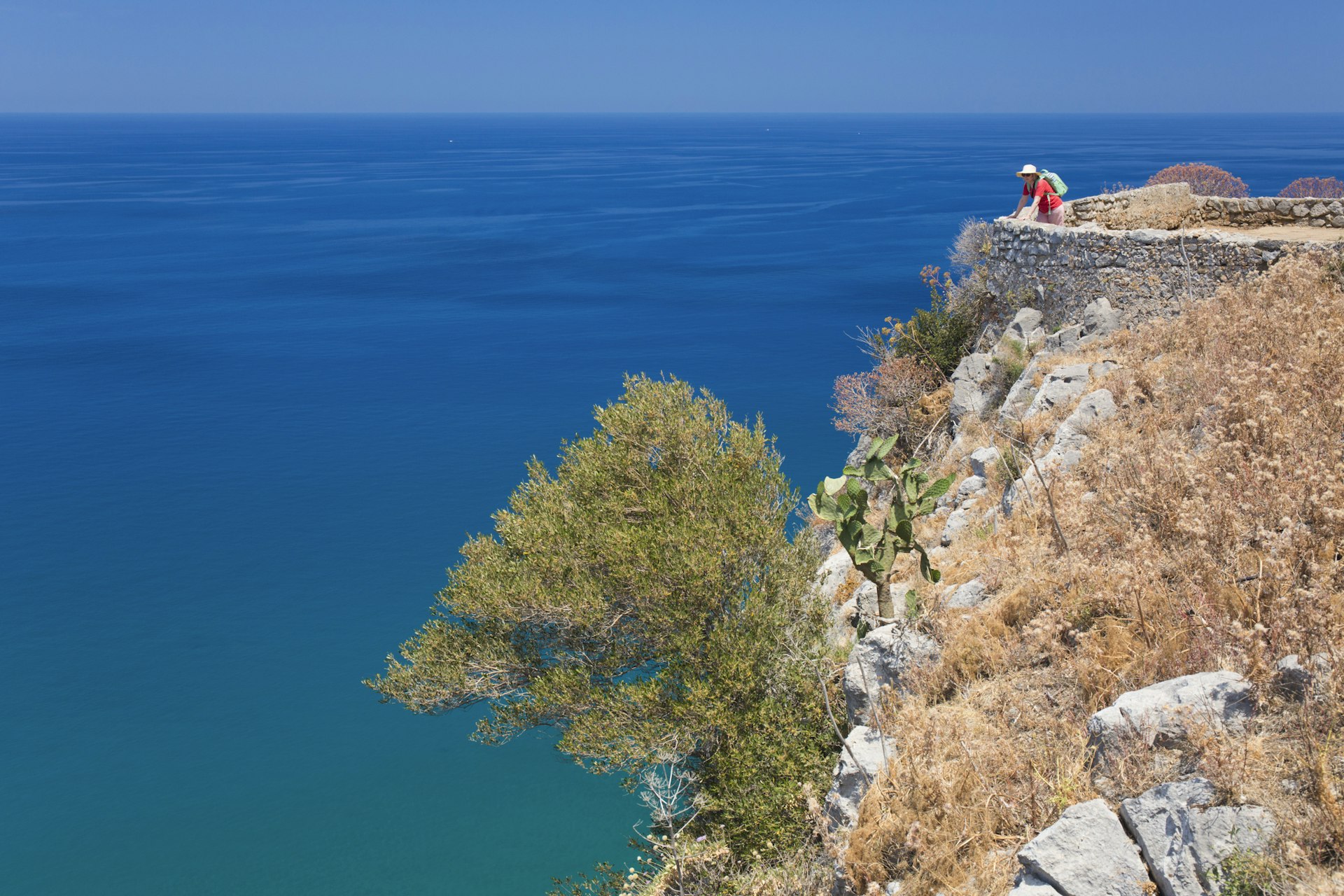 A hiker stands at an ancient stone wall at the top of a cliff admiring the views out to sea