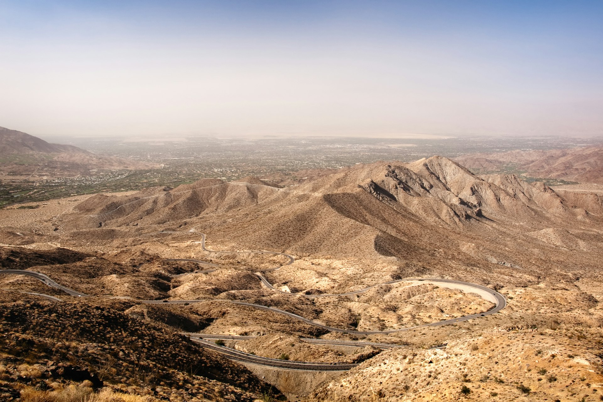 A long road weaves through a mountainous desert landscape with a city in the distance