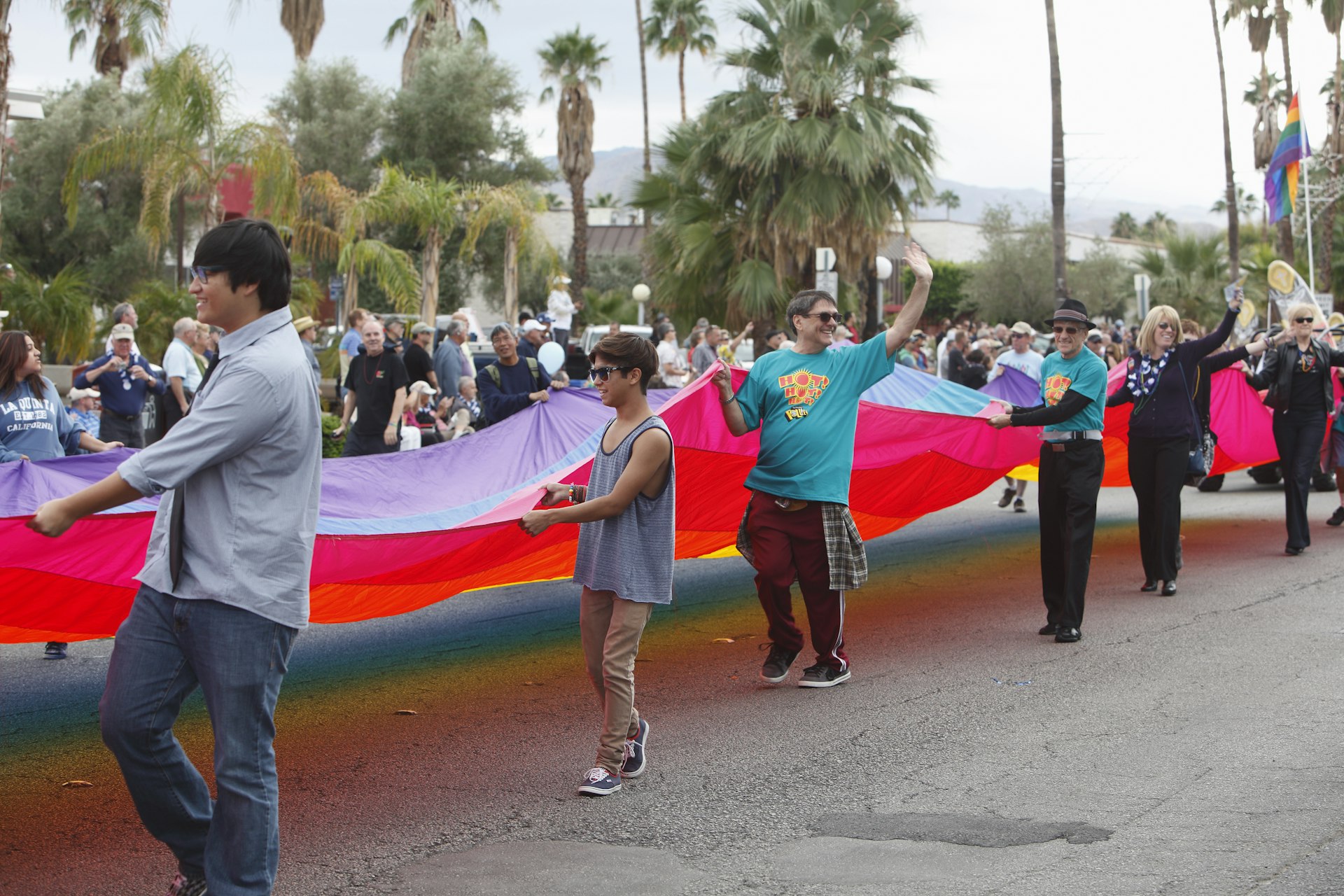 A group of people smile and wave at passers-by as they carry a huge rainbow-striped flag down the street as part of a Pride parade