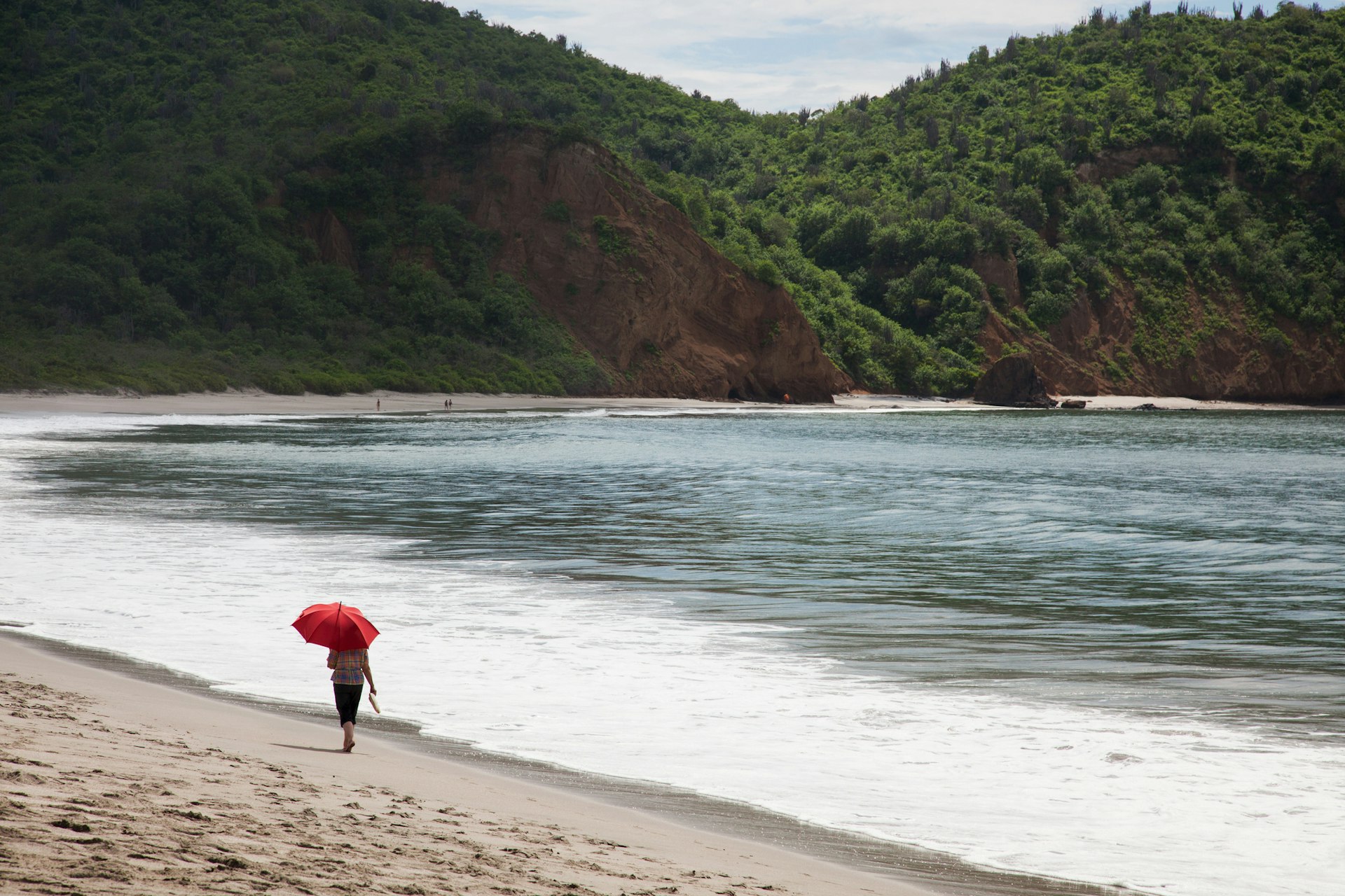 Woman with red umbrella strolls along a beach on a cloudy day