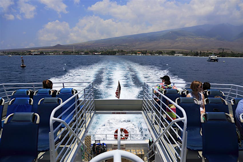 A shot from the back of a ferry with several passengers watching a volcanic island disappear in the distance