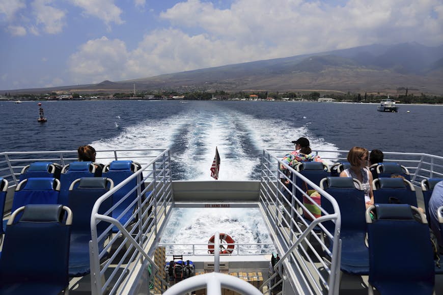 A shot from the back of a ferry with several passengers watching a volcanic island disappear in the distance