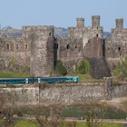 Comwy, Wales - April 9, 2015: Conwy Castle and a Passenger Train North Wales. The railway lone runs along the base of the medeval castle and the Virgin Trains service passes the castle. People can be seen on the catsle walls. The sky is clear and blue.