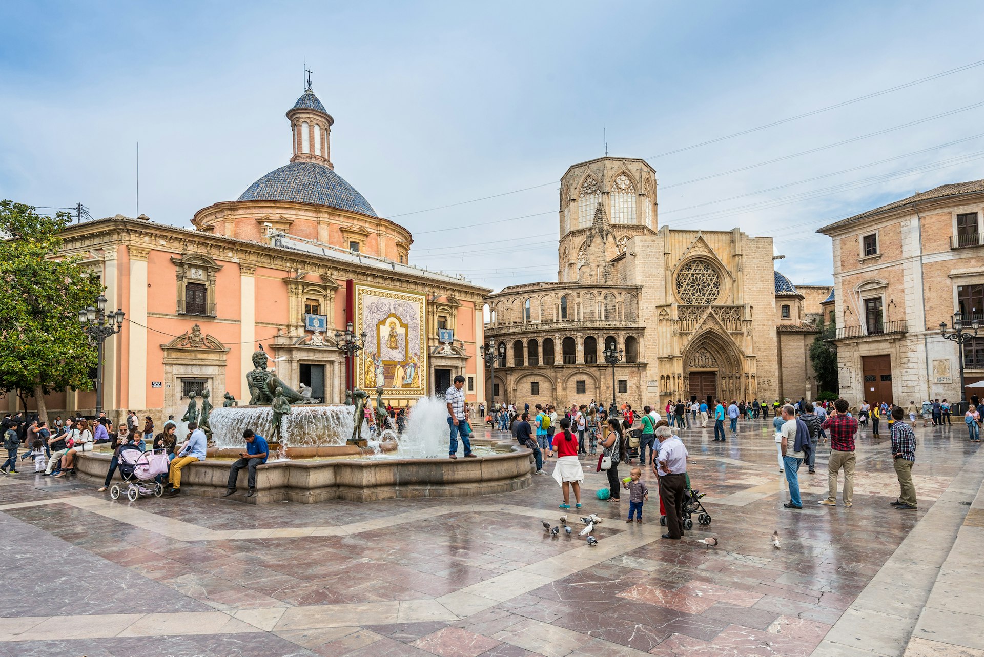 A square lined with historic stone buildings and a large fountain in the center. People are pausing for photos and to enjoy the architecture