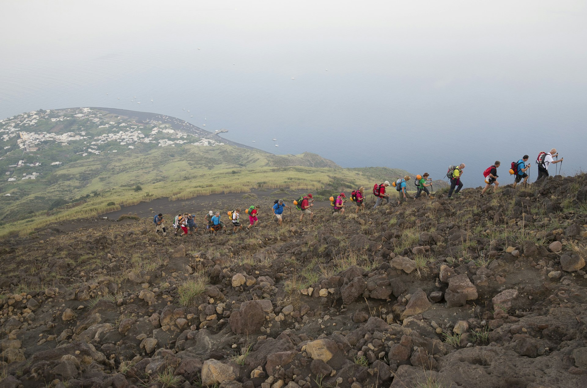 Several hikers in a line following a path up the side of a steep volcano. The town and coastline are in the distance below them