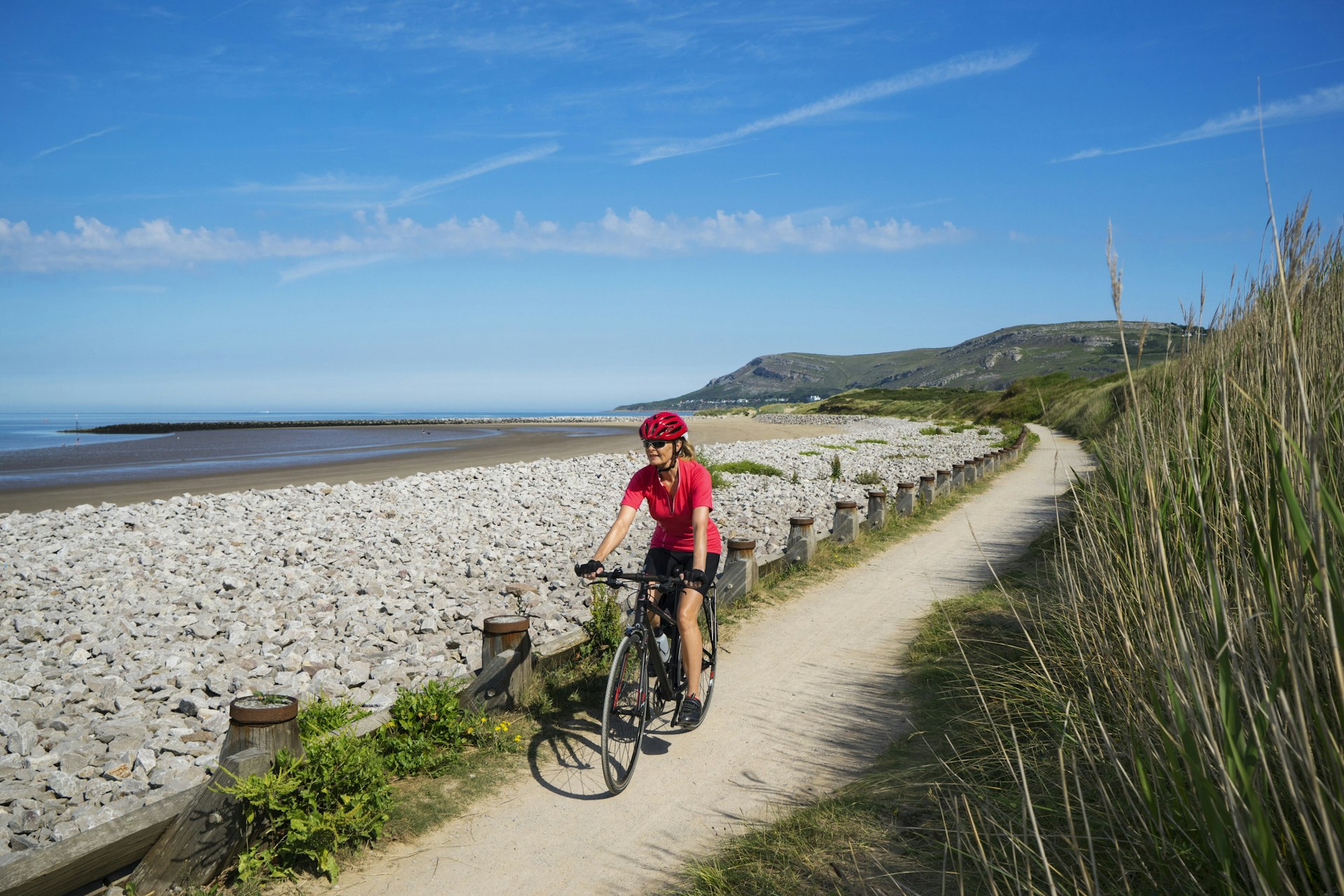 A white female cyclist rides a bike along a paved flat coastal path with a sandy beach