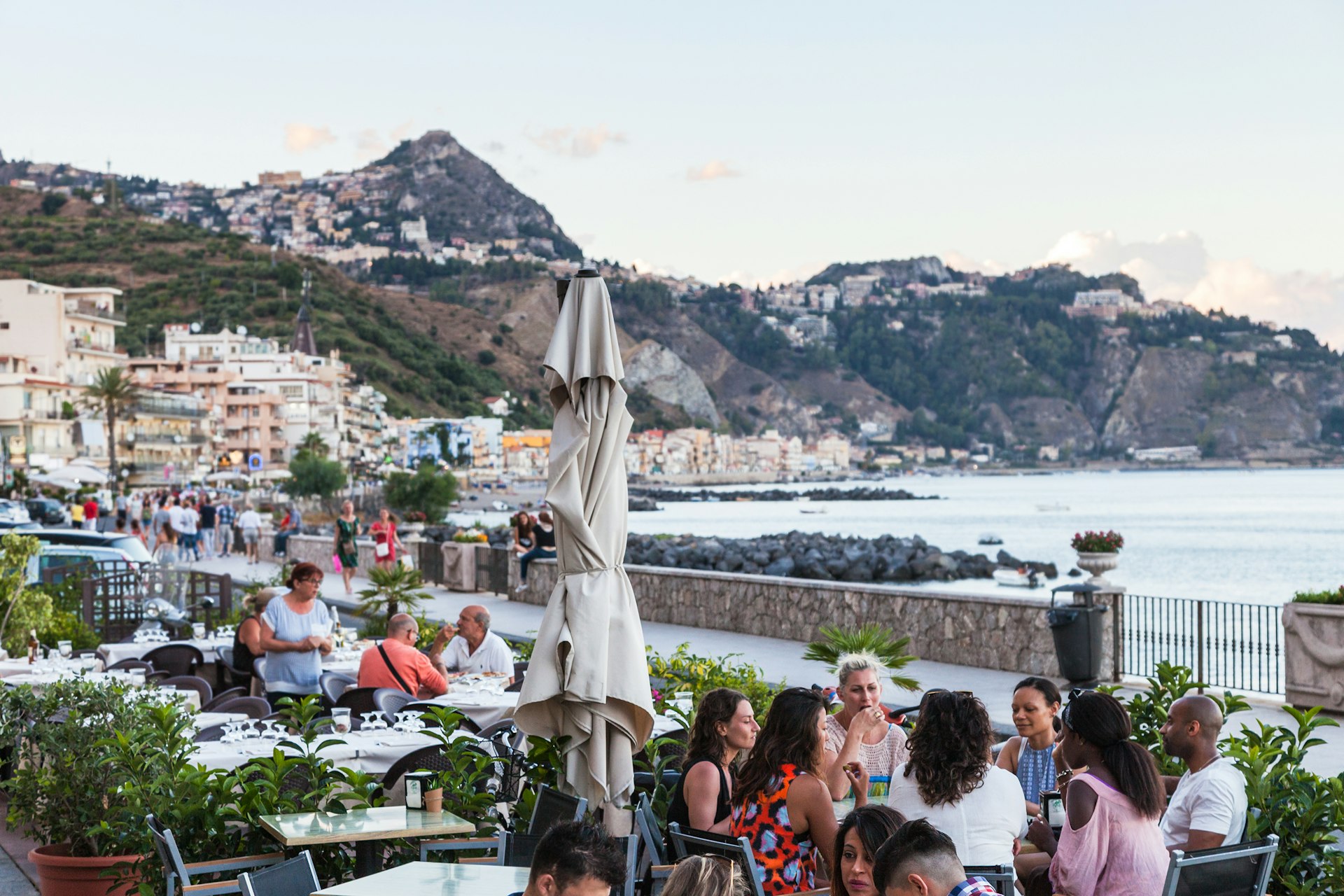 Groups of people sit at restaurant tables on a terrace by the sea 