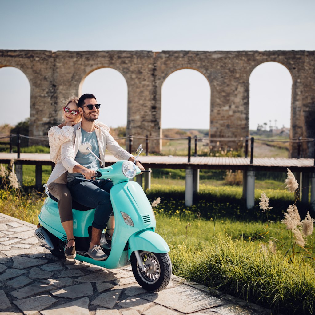 Young loving couple with vintage motorcycle riding next to ancient stone aqueduct monument in Cyprus