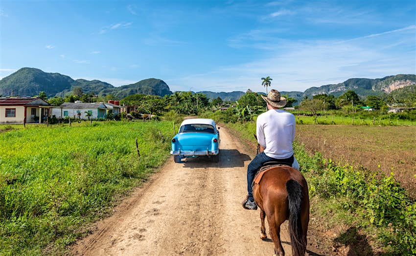 A blue classic car passes a cowboy-hatted man on a horse on a dirt road leading into Vinales, Cuba