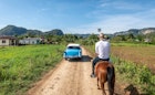 A classic car passes a man on a horse on a dirt road in Vinales.