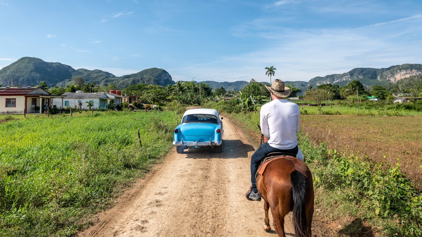 A classic car passes a man on a horse on a dirt road in Vinales.
