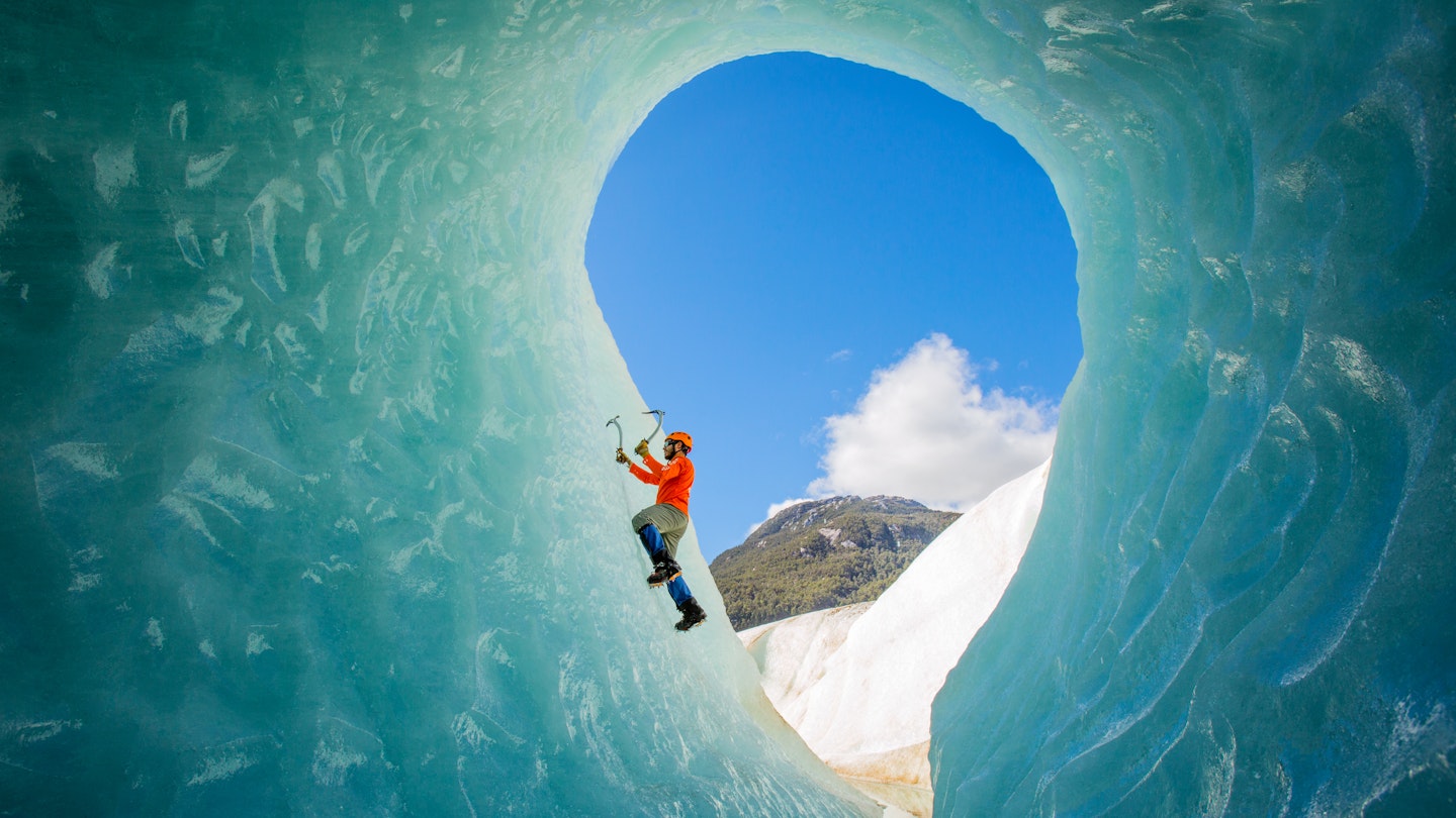 A solo male ice climber working his way up on an ice wall in a glacier. Lifestyle image created from the inside of an ice cave.