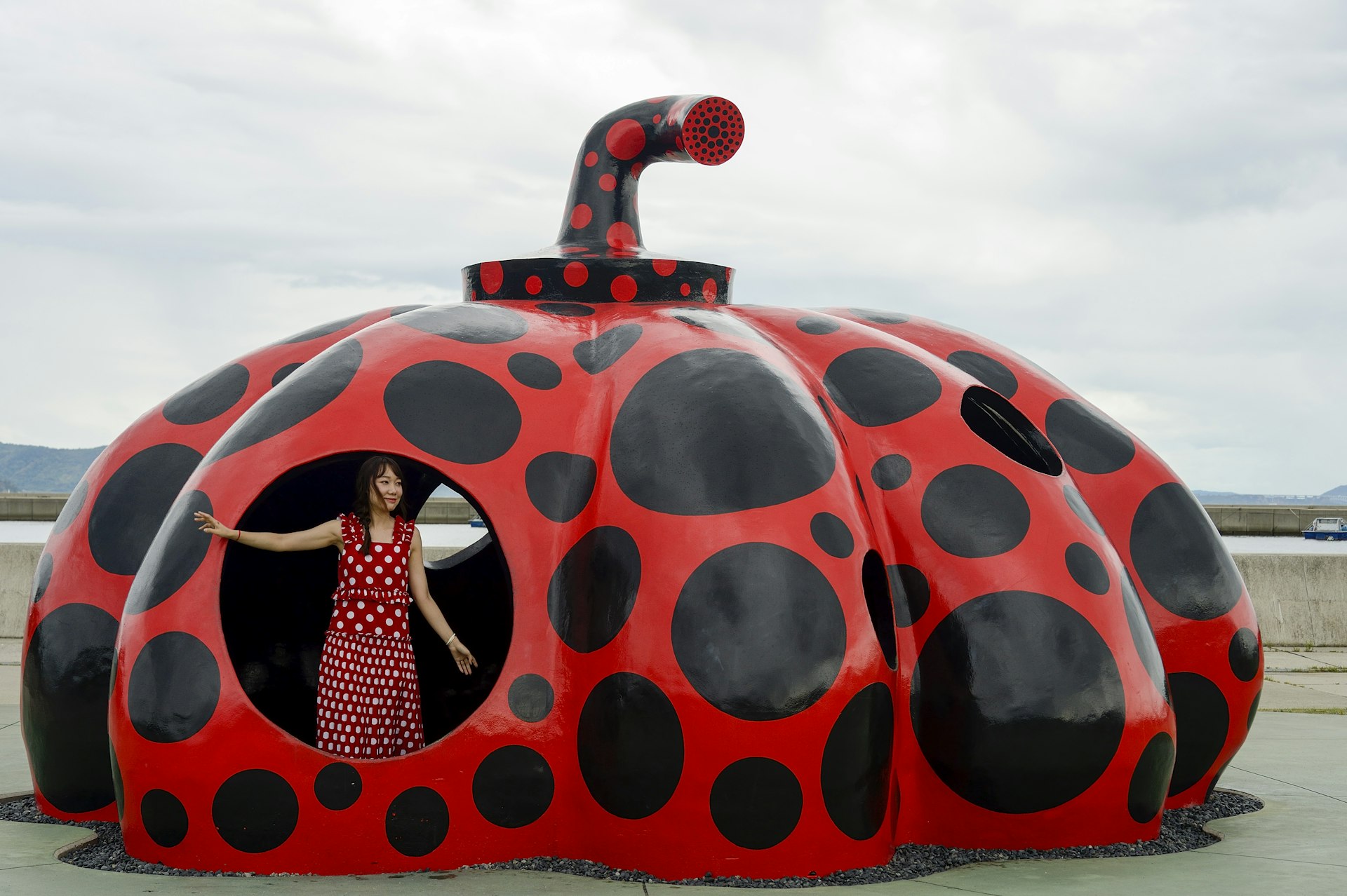 A woman wearing a red and white polka-dot dress stands inside a large red and black polka-dot pumpkin in Naoshima. 