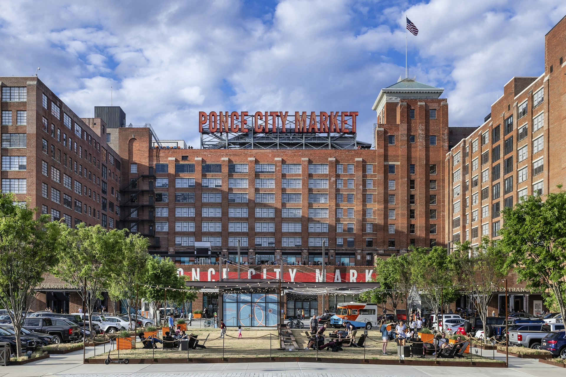 Exterior shot of the brick building of the Ponce city Market. There is a large sign of the market's name at the top of the building. In front there are chairs and tables where people are sitting. 