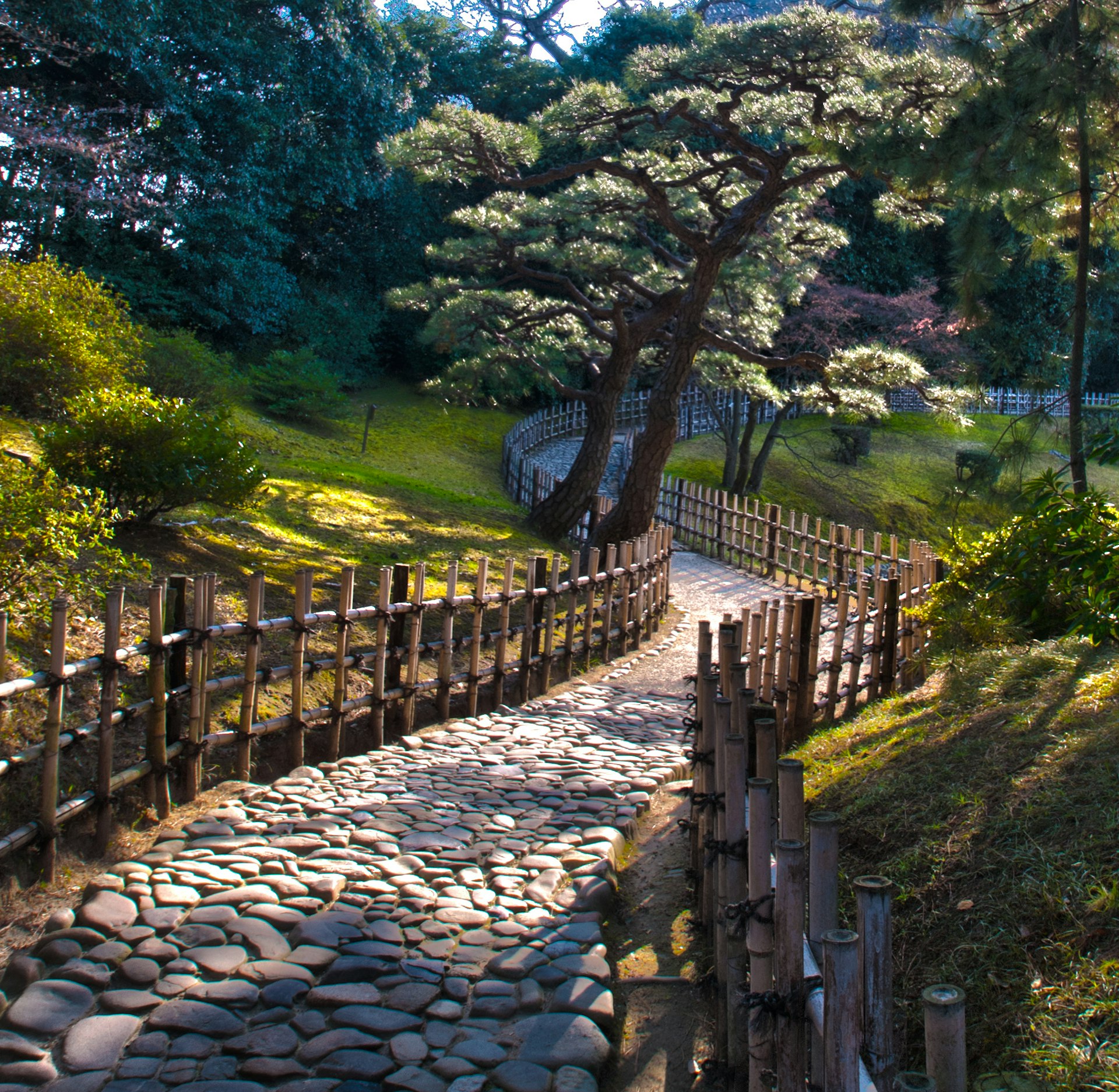 A wooden fence flanks a stone path leading through a serene village at Ritsurin Koen in Takamatsu city in Japan. There are all trees scattered throughout the grassy hills.  