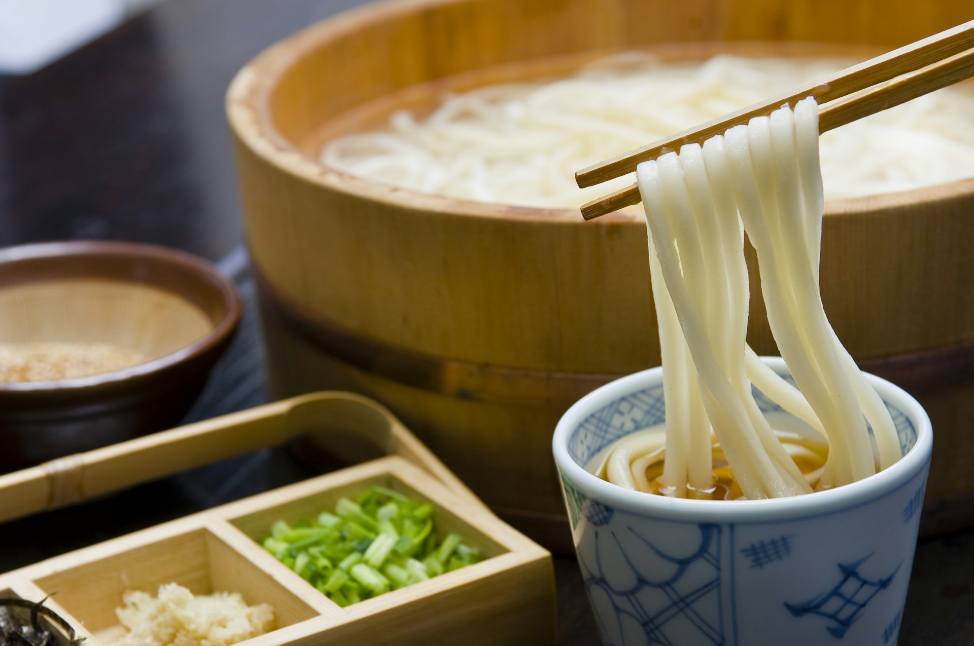 A pair of chopsticks holds some Udon noodles above a small bowl filled with broth. Off to the left is a small wooden box with green onions and seasoning. Off to the right there is a large wooden bowl filled with Udon noodles. 