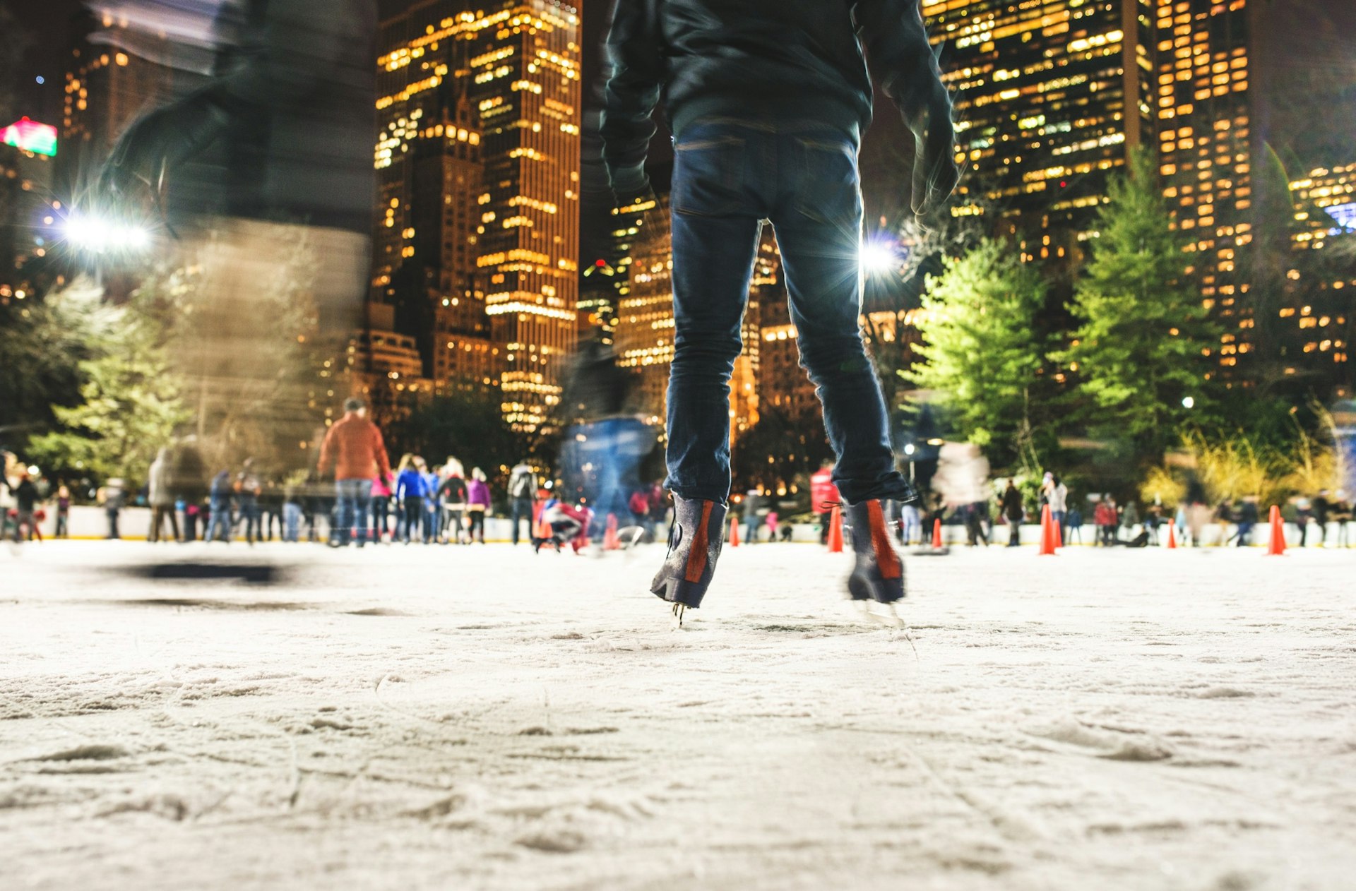 A person wearing skates is the focal point of the frame. In the background there are other people skating at Central Park in night. You can also see tall green trees to the right. 