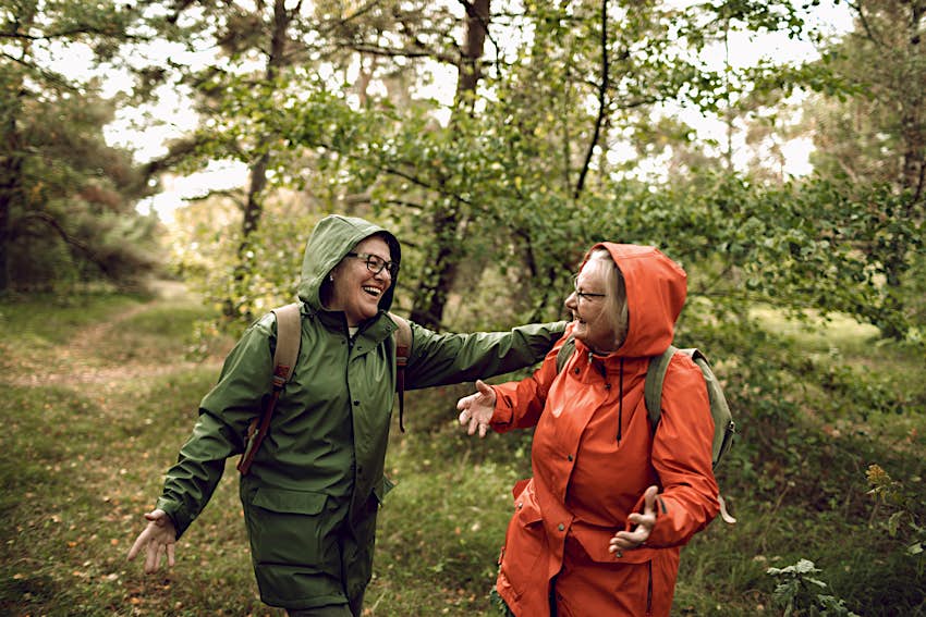 Two women in raincoats walking through a forest and laughing together