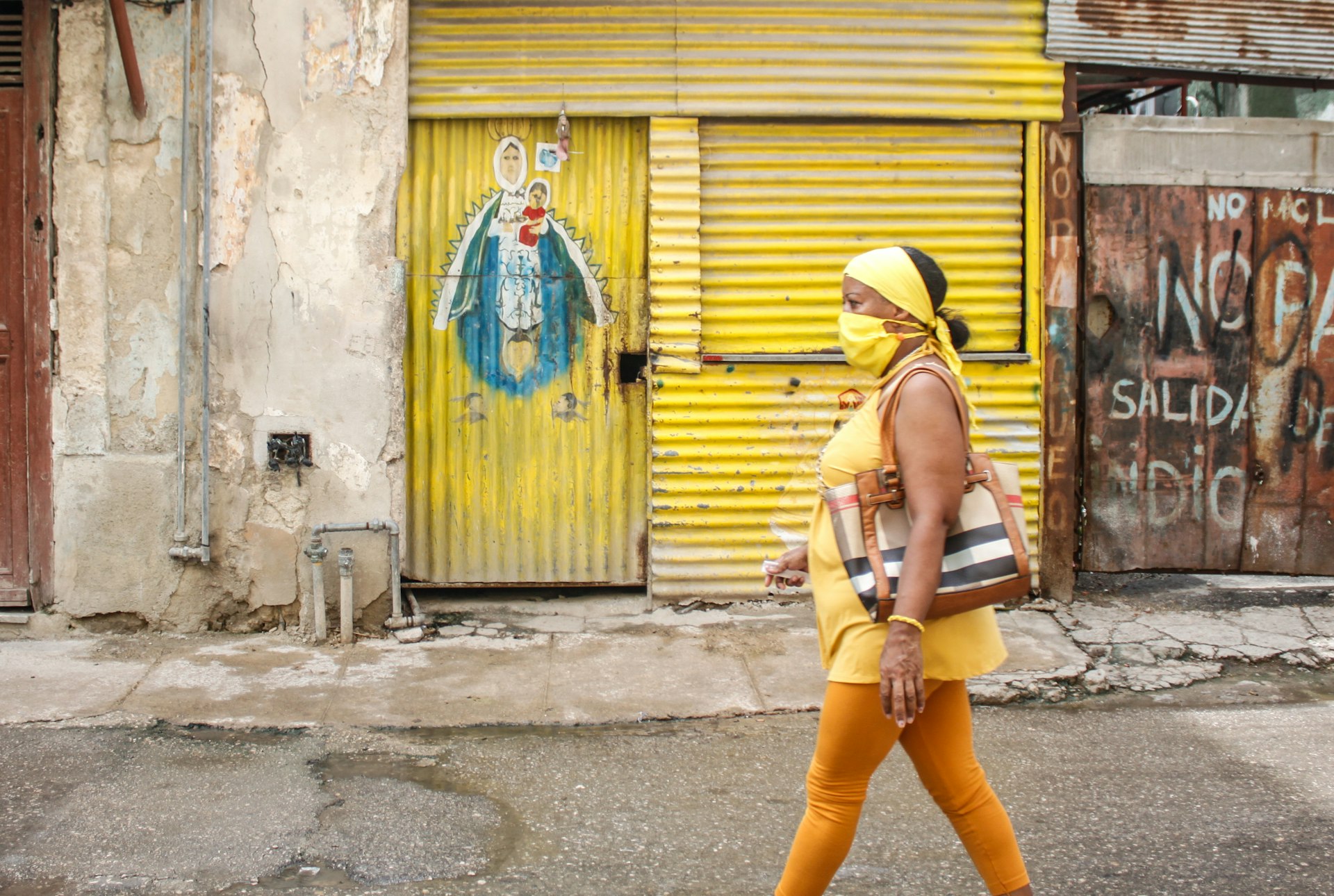 A woman in a yellow outfit with matching mask walks in front of a yellow storefront with street art in Havana