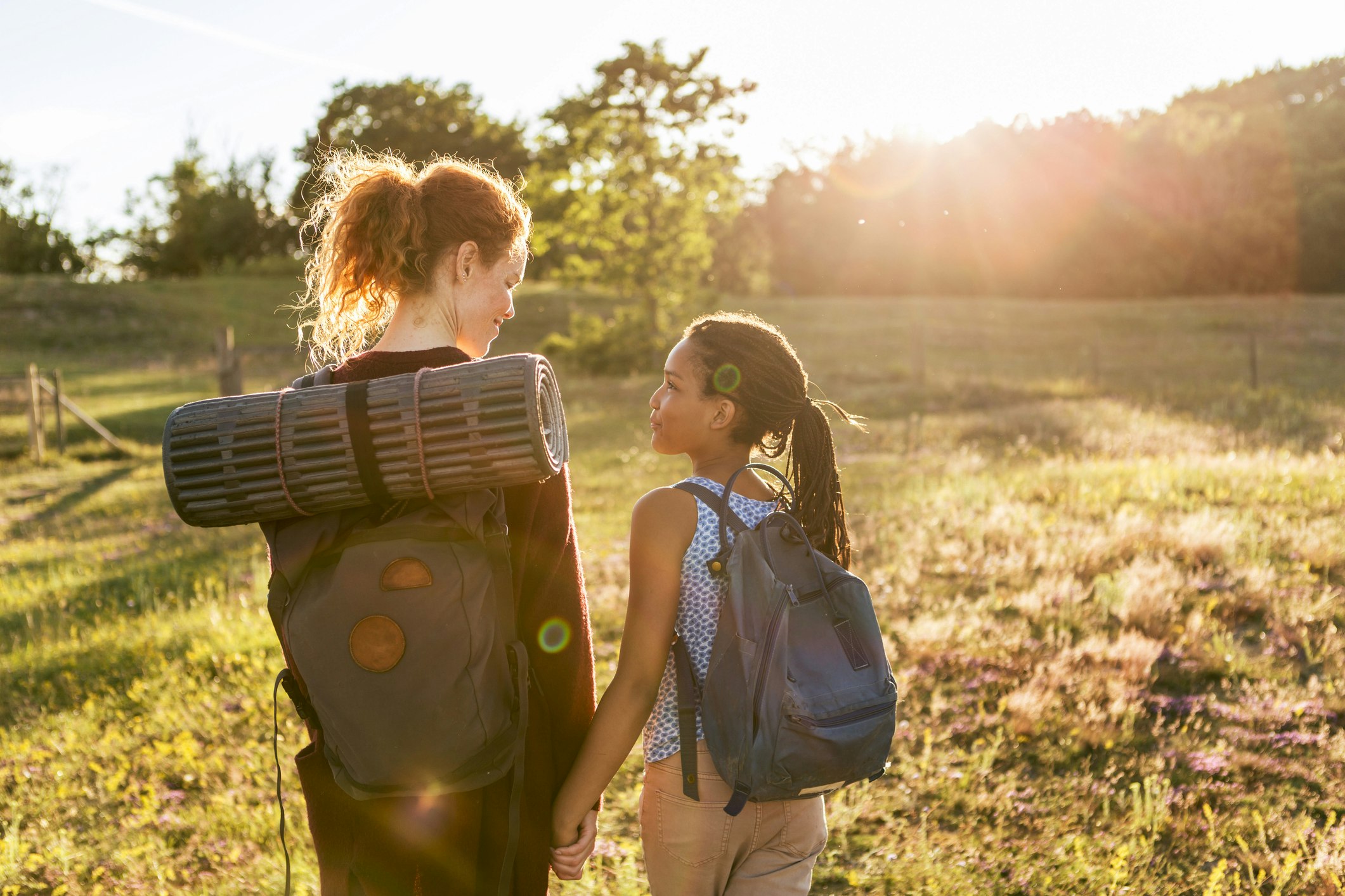 Rear view of woman and girl with backpacks hiking on grass through a park during sunset