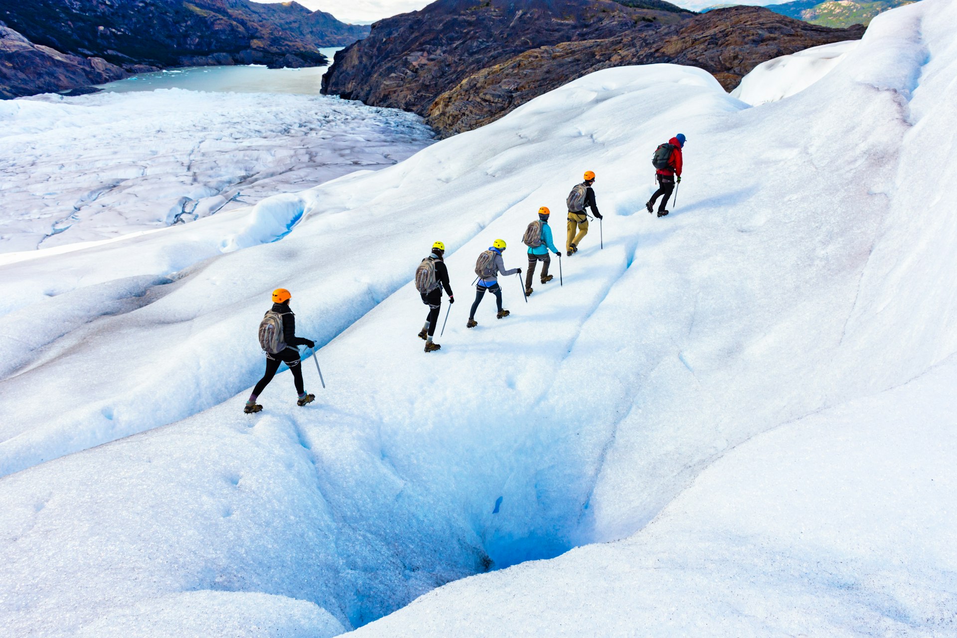 Six trekkers in full hiking gear walk in a line on a glacier