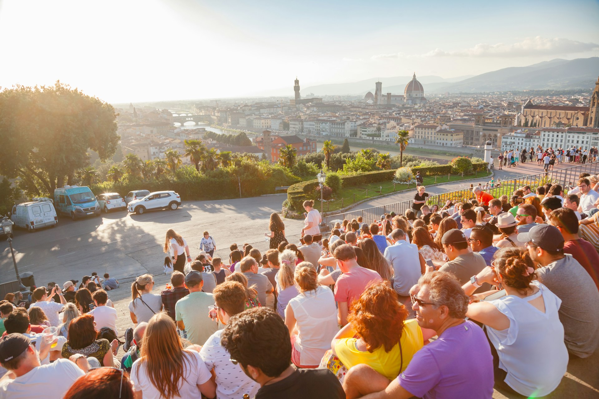 People gatherto enjoy the sunset over a city skyline. A car is blocking in another car nearby