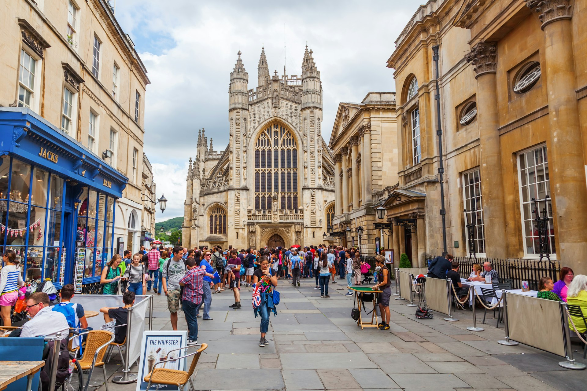 People walk in the street in front of the Bath Abbey, a towering church with a huge arched pane of stained glass facing the plaza.
