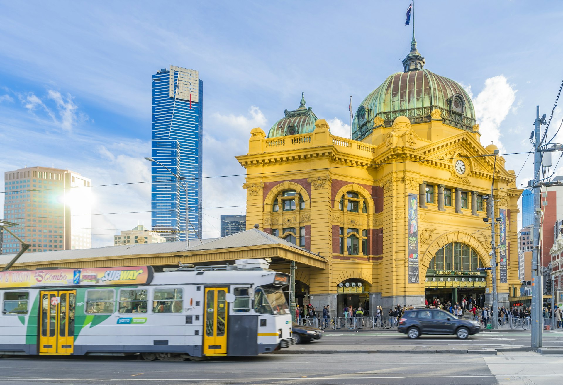 Flinders Street Railway Station in Melbourne with tram, Eureka Tower and other modern buildings near sunset.