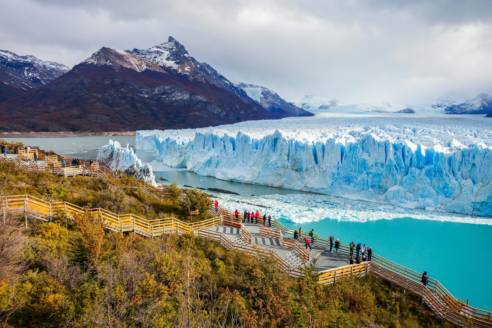 A huge glacier of white ice with a blue tinge stretches out into the distance with rocky peaks either side. A group of tourists admire it from a raised platform