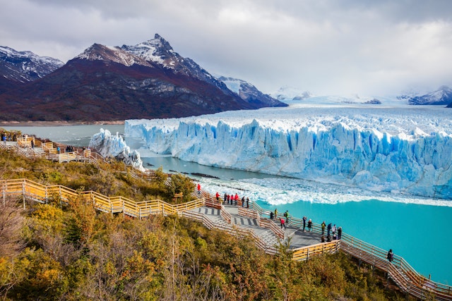 A huge glacier of white ice with a blue tinge stretches out into the distance with rocky peaks either side. A group of tourists admire it from a raised platform