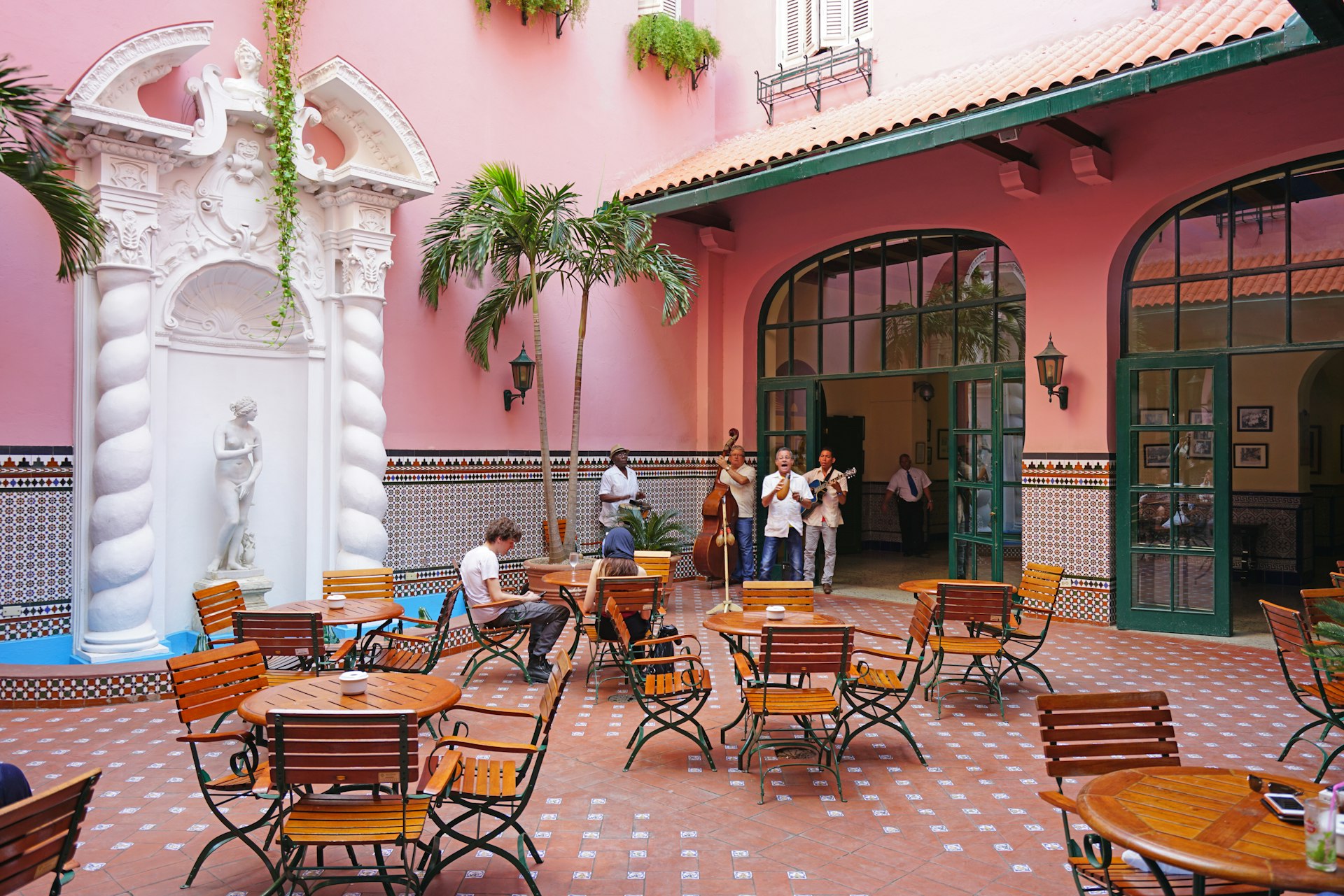 Musicians perform in the corner of a courtyard of the landmark historic Hotel Sevilla Havana, which is painted in bright pink with a couple of onlookers sat at tables looking on