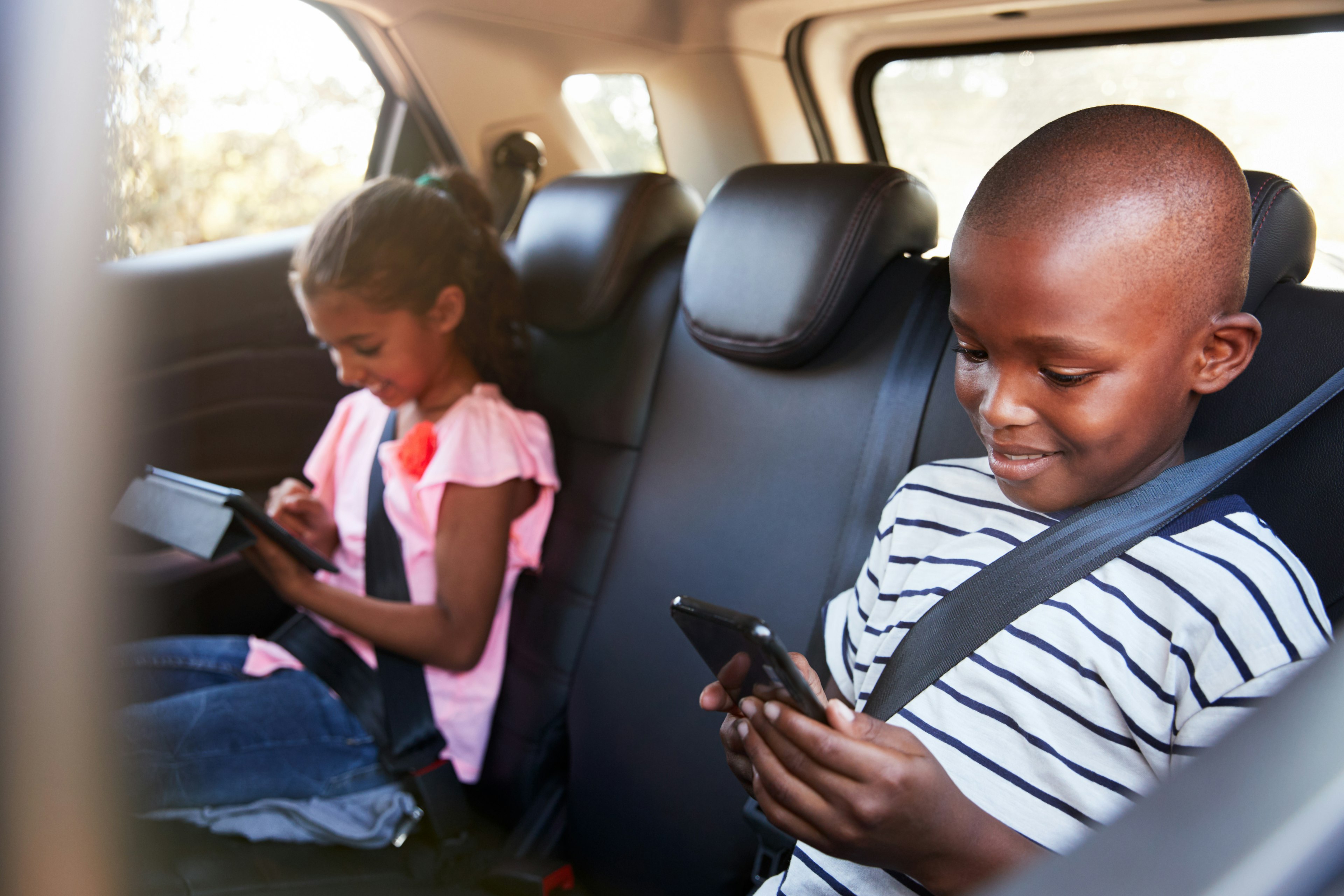 Boy and girl in the back seat of a car using a tablet and smartphone.