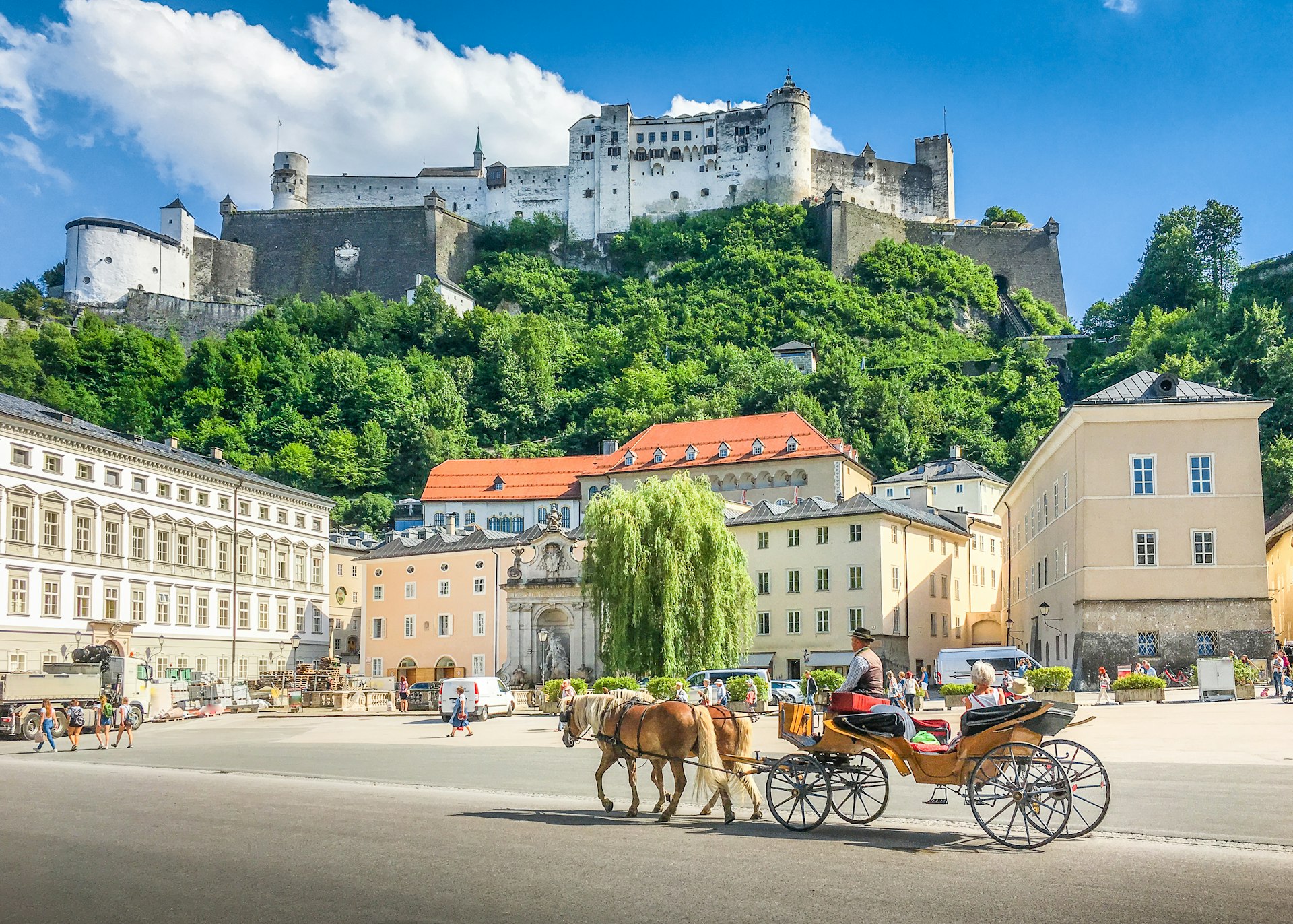 A horse-drawn carriage in the central square of a city. A fortress-like building looms above on a clifftop