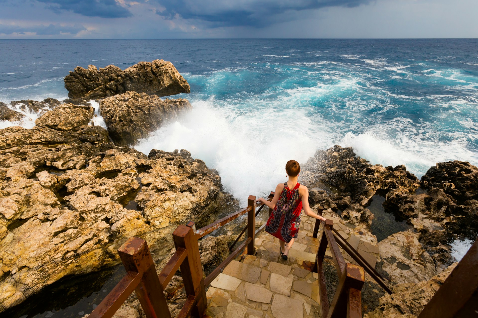 A woman climbs down steps at Cape Greco in front of rough seas