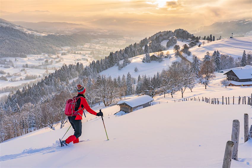 A person in full ski gear snowshoes her way through thick snow in a mountain region as the sun sets