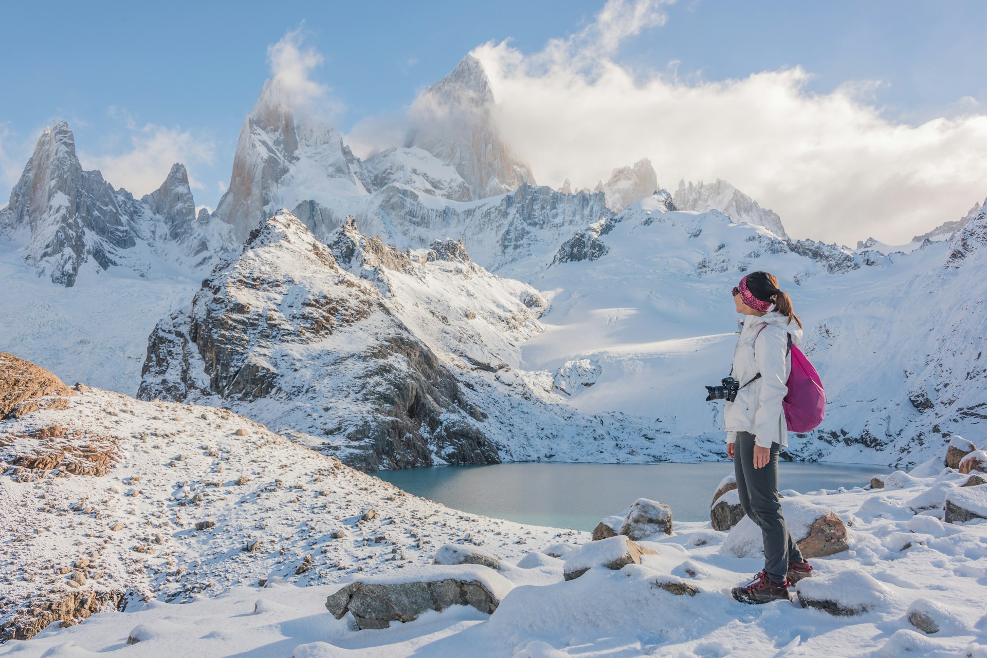 A woman in hiking gear admires the view of a snow-covered mountain range with several distinct jagged peaks